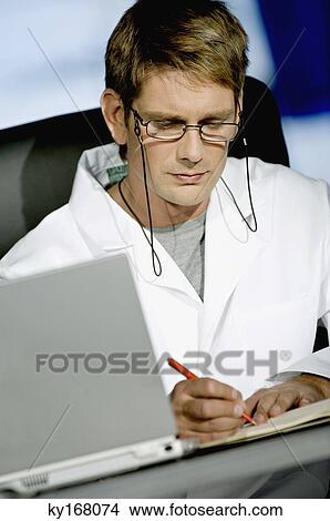 Male doctor sitting at a desk in his office and writing in a notebook View Large Photo Image Picture - Male doctor sitting at a desk in his office and writing in a notebook. Fotosearch