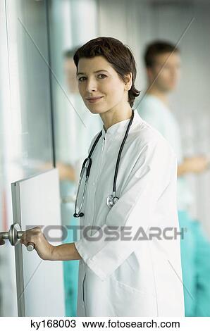 Stock Image - Portrait of a female doctor holding a door handle and smiling. Fotosearch