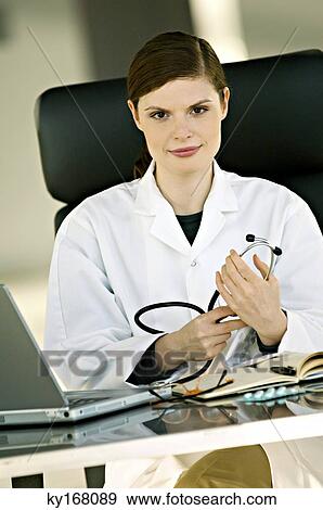 Portrait of a female doctor sitting at a desk and holding a stethoscope in her office View Large Photo Image Stock Photo - Portrait of a female doctor sitting at a desk and holding a stethoscope in her office. Fotosearch