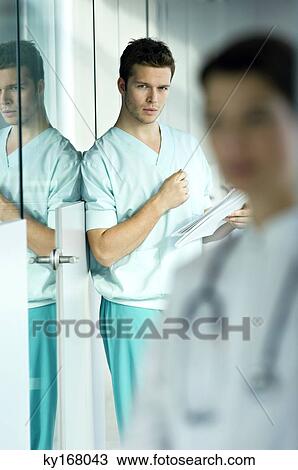 Stock Image - Portrait of a male doctor holding a medical record and leaning against a wall. Fotosearch