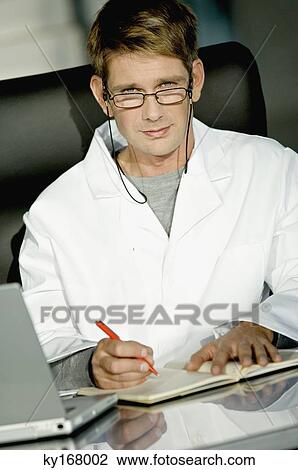 Portrait of a male doctor sitting at a desk in his office and writing in a notebook View Large Photo Image Stock Image - Portrait of a male doctor sitting at a desk in his office and writing in a notebook. Fotosearch