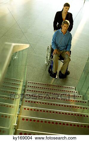 Picture - Male patient sitting in a wheelchair and a mid adult woman standing beside him near a staircase. Fotosearch