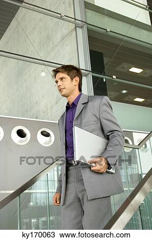 Businessman holding a laptop and walking down a staircase View Large Photo Image Stock Image - Businessman holding a laptop and walking down a staircase. Fotosearch