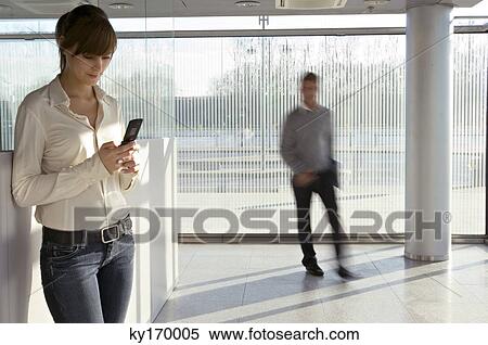 Stock Photography - Businesswoman using a mobile phone with a businessman walking in the background. Fotosearch