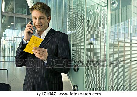 Stock Photo - Businessman holding an airplane ticket and talking on a mobile phone at an airport. Fotosearch