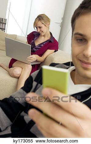 Close-up of a young man holding an MP3 player with a young woman using a laptop behind him View Large Photo Image Stock Photography - Close-up of a young man holding an MP3 player with a young woman using a laptop behind him. Fotosearch