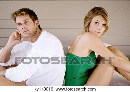 Stock Photograph - Side profile of a mid adult man and a young woman sitting back to back on the bed. Fotosearch