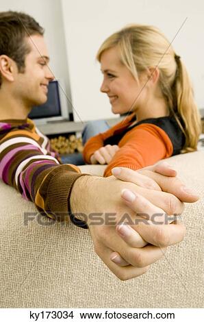 Side profile of a young couple sitting on a couch and holding each other's hands View Large Photo Image Picture - Side profile of a young couple sitting on a couch and holding each other's hands. Fotosearch