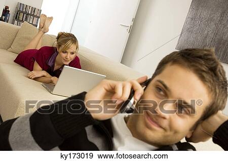Young man using a mobile phone with a young woman using a laptop in the background View Large Photo Image Stock Photo - Young man using a mobile phone with a young woman using a laptop in the background. Fotosearch
