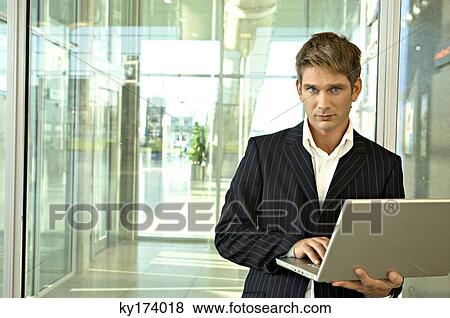 Businessman standing and using a laptop at an airport View Large Photo Image Stock Photo - Businessman standing and using a laptop at an airport. Fotosearch