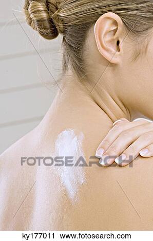 Stock Image - Rear view of a young woman applying moisturizer on her shoulder. Fotosearch