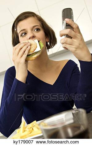 Close-up of a young woman eating a burger and looking at a mobile phone View Large Photo Image Stock Photo - Close-up of a young woman eating a burger and looking at a mobile phone. Fotosearch