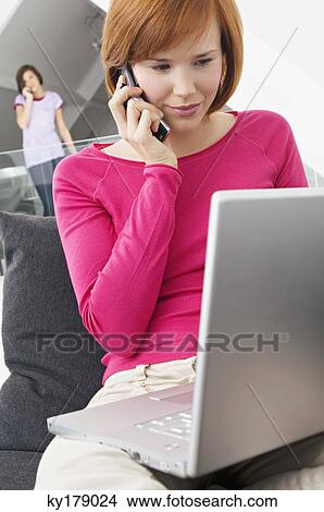Picture - Close-up of a young woman using a laptop and talking on a mobile phone. Fotosearch