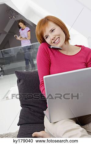 Stock Image - Young woman using a laptop and talking on a mobile phone. Fotosearch