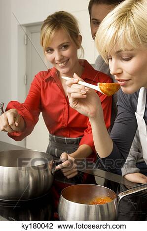 Stock Image - Two young women and a young man preparing food in the kitchen. Fotosearch