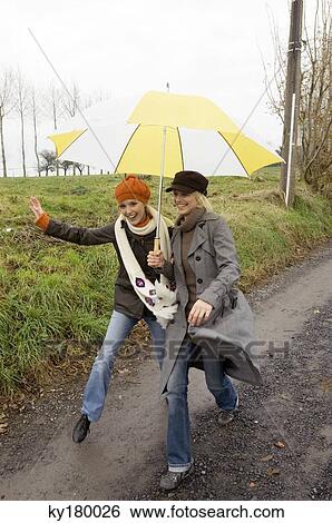 Stock Photograph - Two young women sheltering under an umbrella and walking on a dirt road. Fotosearch
