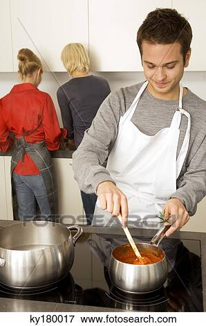 Young man cooking food and two young women standing behind him in the kitchen View Large Photo Image Stock Photo - Young man cooking food and two young women standing behind him in the kitchen. Fotosearch