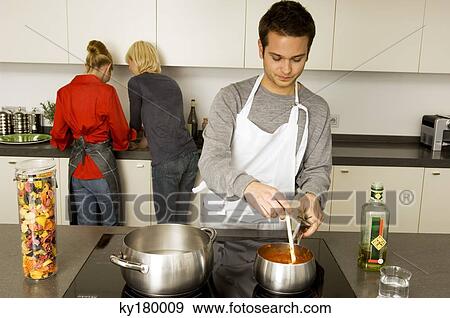 Young man cooking food with two young women standing behind him View Large Photo Image Stock Photo - Young man cooking food with two young women standing behind him. Fotosearch