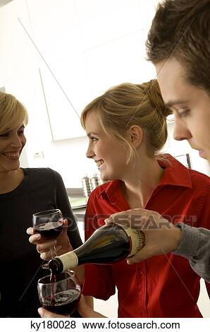 Young man pouring wine into a wine glass with two young women smiling beside him View Large Photo Image Stock Photo - Young man pouring wine into a wine glass with two young women smiling beside him. Fotosearch