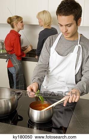 Young man preparing food with two young women talking with each other in the background View Large Photo Image Stock Image - Young man preparing food with two young women talking with each other in the background. Fotosearch