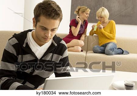 Young man using a laptop with two young women listening to an MP3 player in the background View Large Photo Image Picture - Young man using a laptop with two young women listening to an MP3 player in the background. Fotosearch
