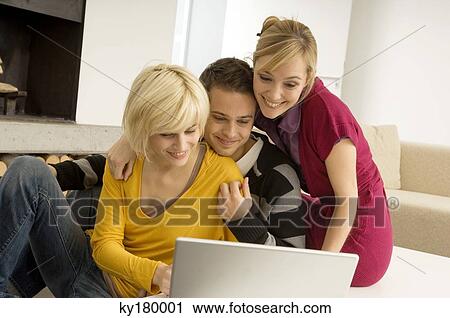Stock Image - Young man with two young women looking at a laptop and smiling. Fotosearch
