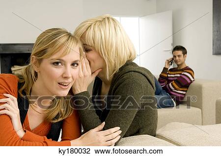 Young woman whispering to her friend with a young man sitting in the background View Large Photo Image Stock Image - Young woman whispering to her friend with a young man sitting in the background. Fotosearch