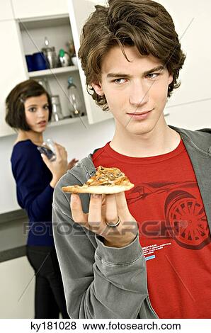 Close-up of a teenage boy holding a slice of pizza and a young woman standing behind him View Large Photo Image Stock Photo - Close-up of a teenage boy holding a slice of pizza and a young woman standing behind him. Fotosearch