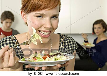 Portrait of a young woman eating salad with her friends in background View Large Photo Image Stock Image - Portrait of a young woman eating salad with her friends in background. Fotosearch