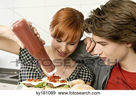 Young woman pouring tomato sauce on burgers with a teenage boy standing beside her View Large Photo Image Stock Photo - Young woman pouring tomato sauce on burgers with a teenage boy standing beside her. Fotosearch