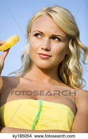 Picture - Portrait of a young blond woman eating pineapple slice. Fotosearch