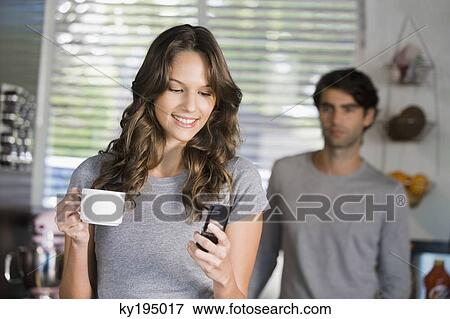 Stock Photo - Woman text messaging with a man standing in the background. Fotosearch