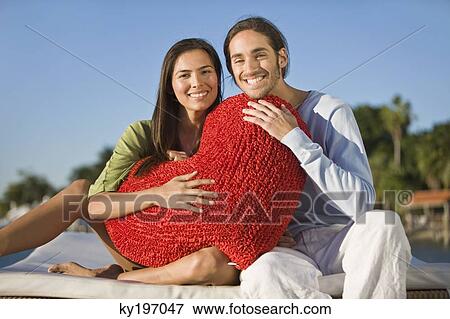 Portrait of a couple holding a heart shaped cushion View Large Photo Image Stock Photo - Portrait of a couple holding a heart shaped cushion. Fotosearch