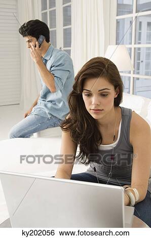 Woman working on a laptop with a man talking on a mobile phone in the background View Large Photo Image Stock Photograph - Woman working on a laptop with a man talking on a mobile phone in the background. Fotosearch