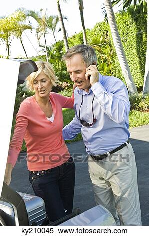 Stock Photography - Man talking on a mobile phone beside his wife loading a luggage into car. Fotosearch