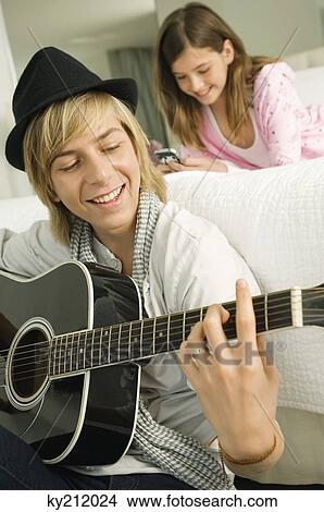 Teenage boy playing a guitar and his sister using a mobile phone behind him View Large Photo Image Picture - Teenage boy playing a guitar and his sister using a mobile phone behind him. Fotosearch