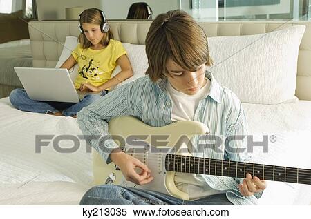Boy playing a guitar and girl working on a laptop behind him View Large Photo Image Stock Photography - Boy playing a guitar and girl working on a laptop behind him. Fotosearch