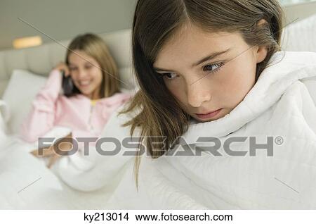 Picture - Close-up of a girl wrapped in a blanket with her sister talking on mobile phone in the background. Fotosearch