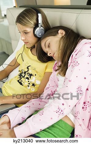 Girl working on a laptop and listening to headphones and her sister sleeping on her shoulders View Large Photo Image Stock Image - Girl working on a laptop and listening to headphones and her sister sleeping on her shoulders. Fotosearch