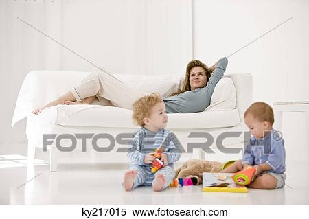 Stock Photography - Babies playing with toys and their mother resting on a couch. Fotosearch