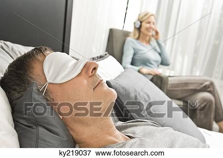 Stock Photo - Man wearing an eye mask and his wife listening to music in the background. Fotosearch