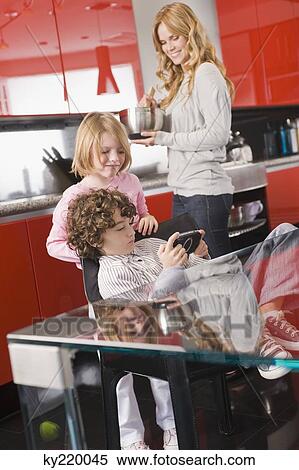 Boy playing a video game with his sister and mother in the background View Large Photo Image Stock Photography - Boy playing a video game with his sister and mother in the background. Fotosearch