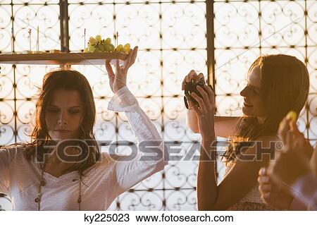 Stock Image - Woman taking a picture of her friend carrying a fruit tray. Fotosearch