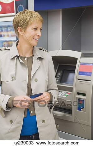 Stock Image - Woman holding a credit card and standing in front of an ATM. Fotosearch