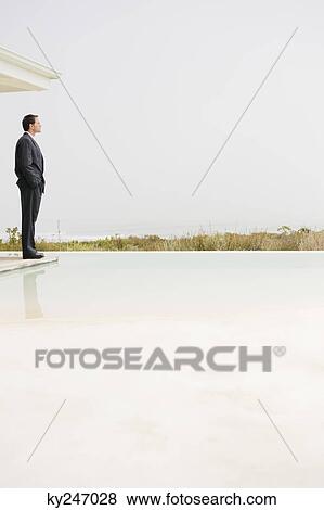Stock Photo - Businessman standing at the poolside and looking at sea view. Fotosearch