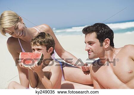 Stock Photography - Boy eating a watermelon with his parents on the beach. Fotosearch