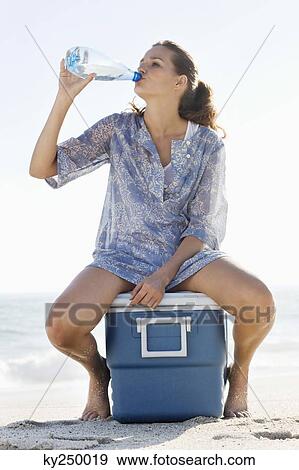 Stock Photo - Woman sitting on an ice box and drinking water on the beach. Fotosearch