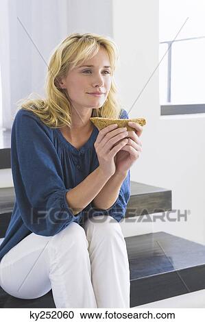Stock Image - Woman sitting on steps and holding a sandwich. Fotosearch