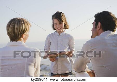Girl reading a book in front of her parents View Large Photo Image Stock Image - Girl reading a book in front of her parents. Fotosearch