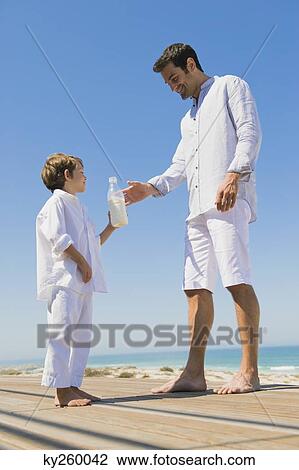 Stock Image - Man giving milk bottle to his son on the beach. Fotosearch